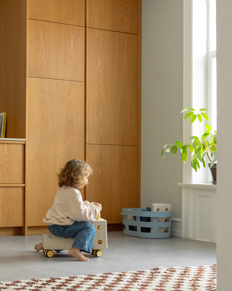 Toddler riding a MODU ride-on toy around the kitchen. Built from Curiosity Set in Sand Grey Honey Yellow.