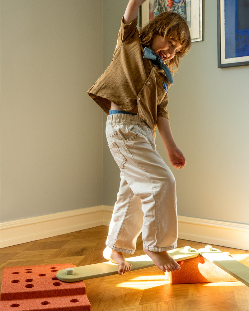 A child walks barefoot across a balance track made with dusty green MODU Balance Beams and burnt orange MODU foam blocks in a living room.