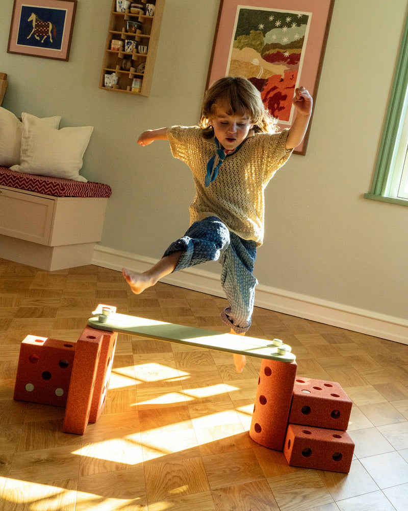 Child jumps over a raised balance beam structure built from burnt orange MODU building blocks and dusty green MODU Balance Beams during indoor play.