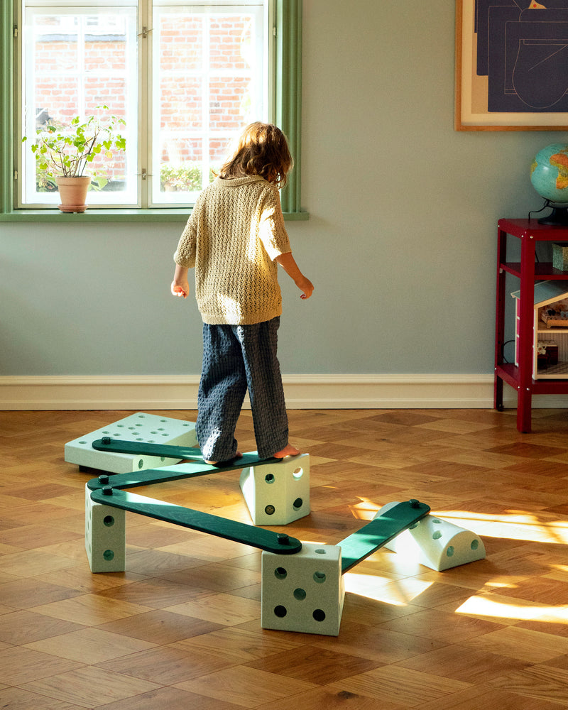 A child walks barefoot along a zig-zag balance track built with forest green MODU Balance Beams and ocean mint MODU foam blocks.