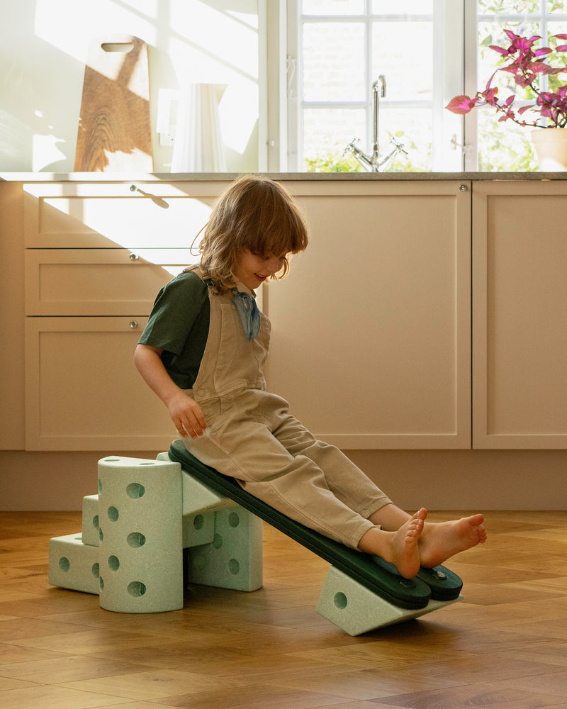 A child slides down a ramp structure built with forest green MODU Balance Beams and ocean mint MODU foam blocks in a bright kitchen.