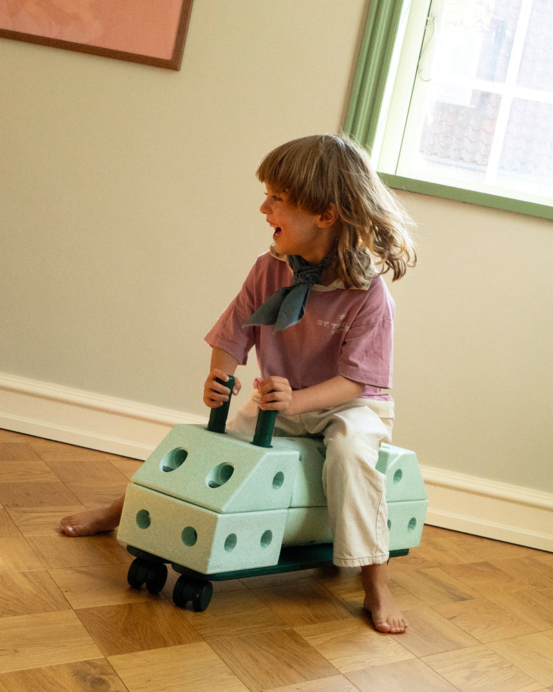 A laughing child scoots across living room on a ride-on toy built from ocean mint MODU building blocks featuring forrest green MODU balance beams.