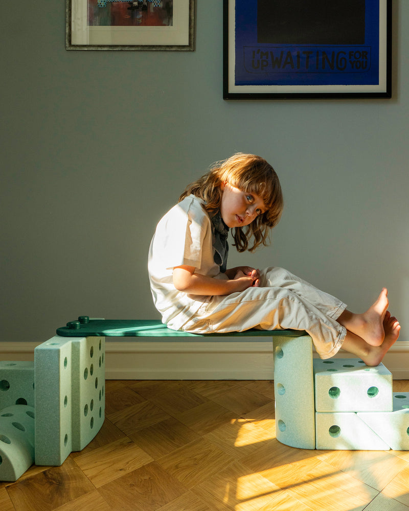 A child sits relaxed on a low bench configuration built from forest green MODU Balance Beams and ocean mint MODU foam blocks.