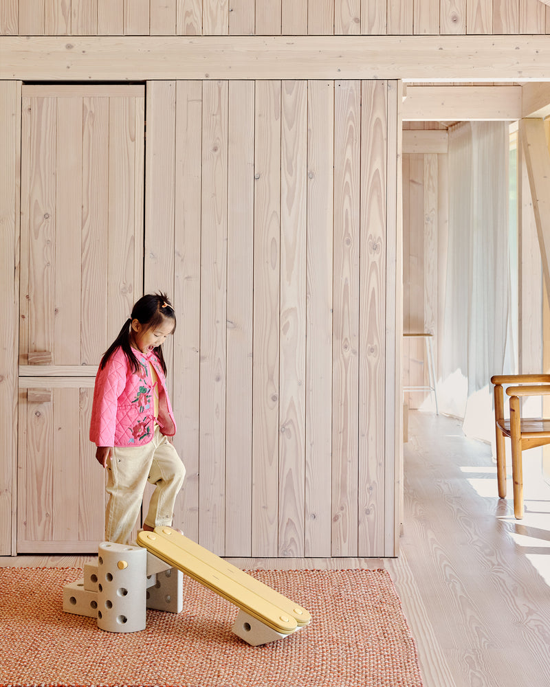 A young girl smiles as she steps down a ramp built from honey yellow MODU Balance Beams and sand grey MODU building blocks.