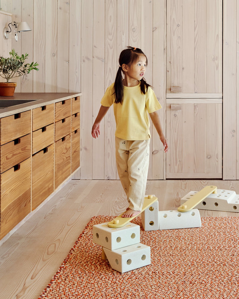 A child walks barefoot along an indoor balance track built with honey yellow MODU Balance Beams and sand grey MODU building blocks.