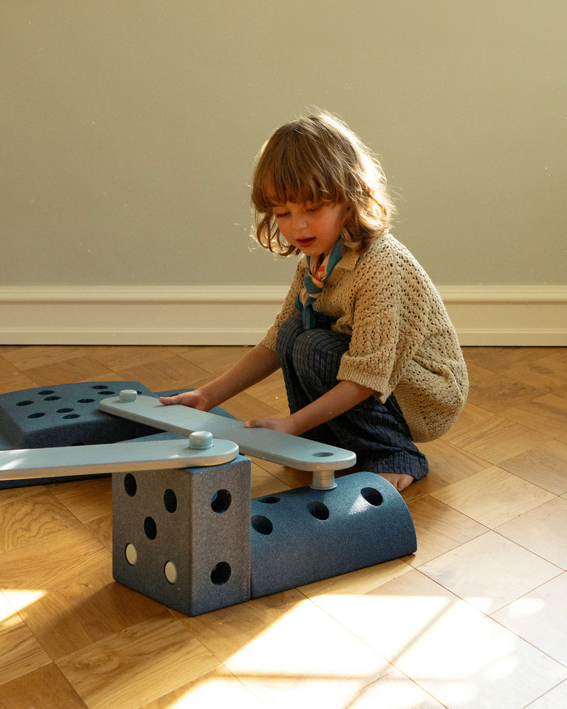 A child crouches down on a wooden floor to build an obstacle course using a sky blue MODU Balance Beam.