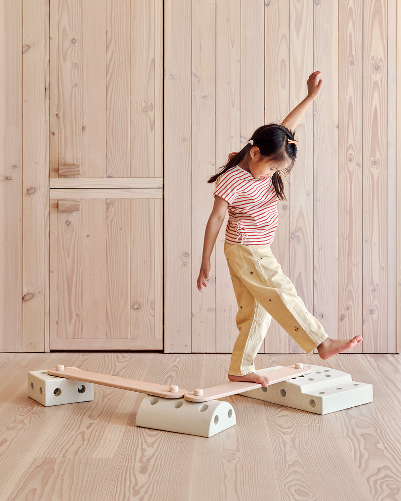A young girl stretches her arm out for balance while walking across a low balance track built with soft rose MODU Balance Beams.