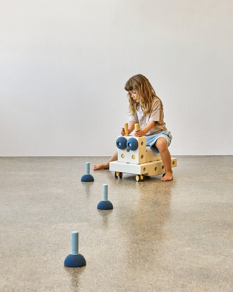 A toddler rides a creative ride-on built from MODU foam blocks, steering through an indoor obstacle course of deep blue MODU Foam Balls used as cones.