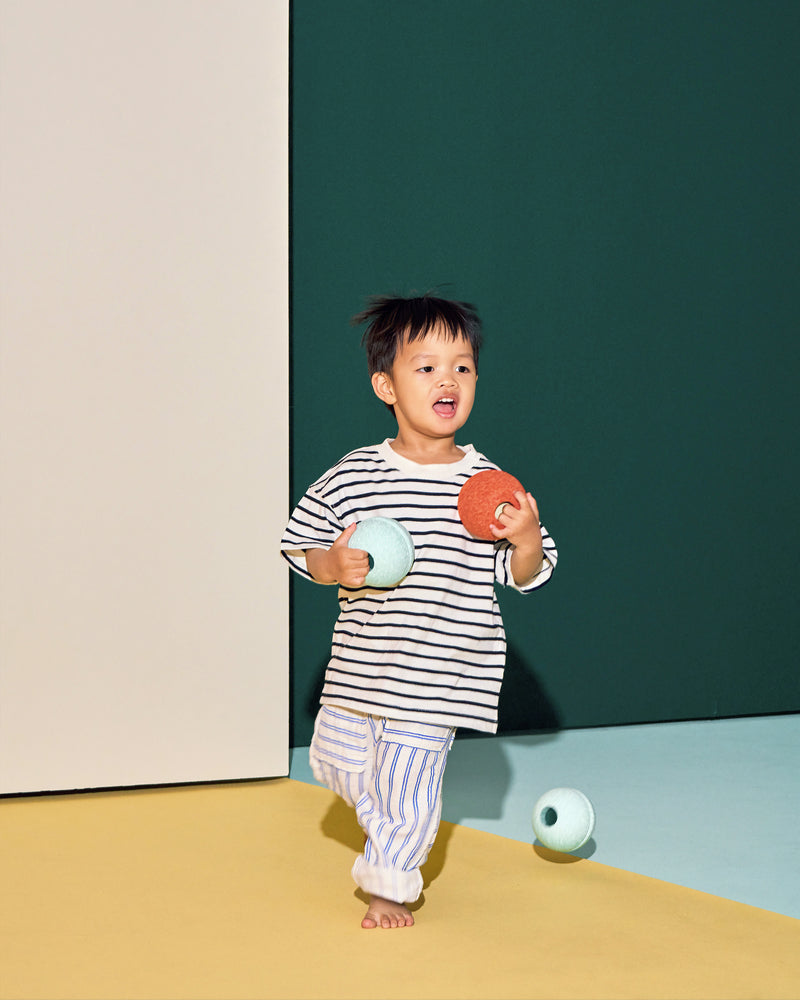 A toddler happily runs across a play area while holding ocean mint and burnt orange MODU Foam Balls during active play.