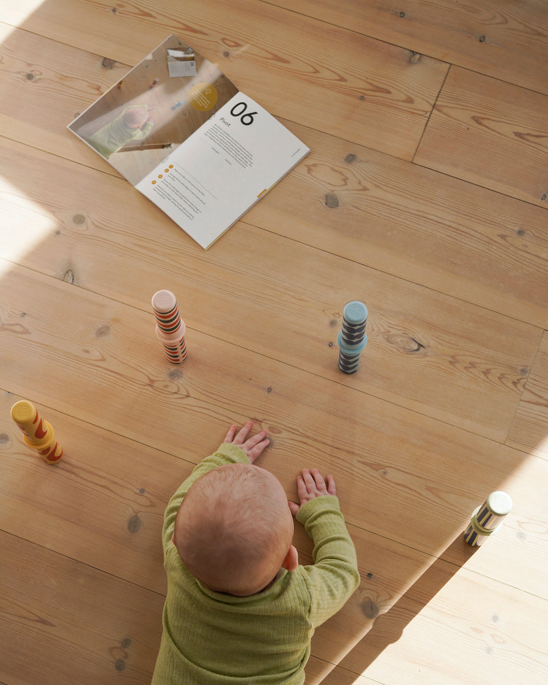 A baby practices pivoting on a wooden floor, reaching for MODU Rattle Pegs stacked in a half circle on the floor.