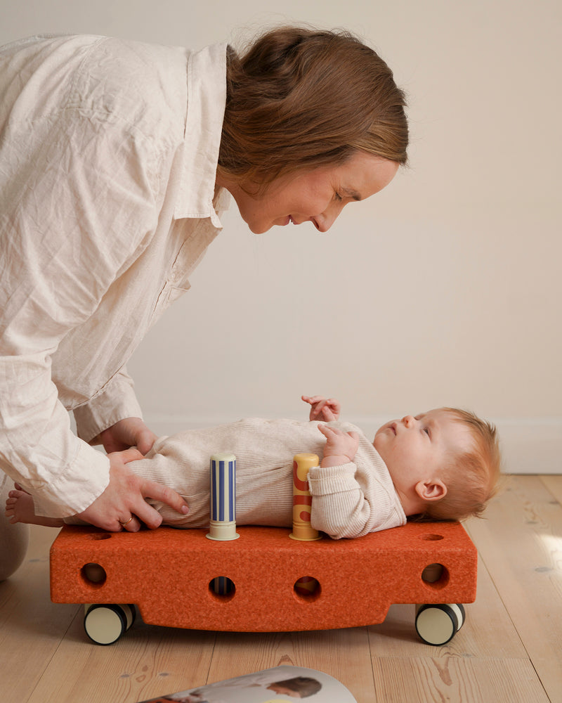 An adult interacts with a baby lying on its back on a scooter board built from the burnt orange MODU Baby Activity Set.