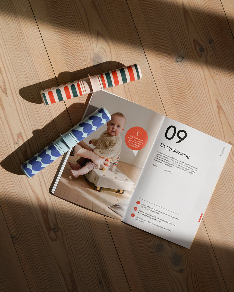 An open MODU activity guide showcasing early development milestone play sits on a wooden floor alongside colorful patterned rattle pegs.