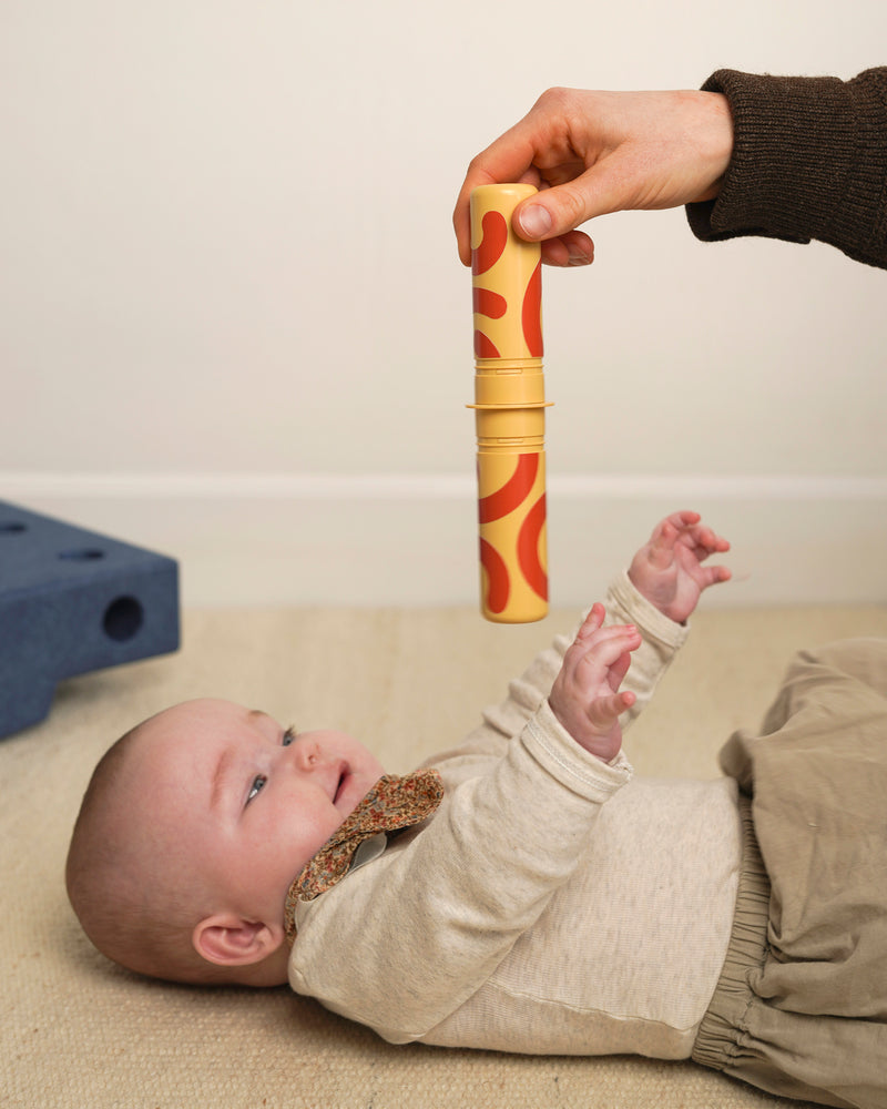 An adult holds a patterned MODU rattle peg above a baby lying down, encouraging active reaching play.