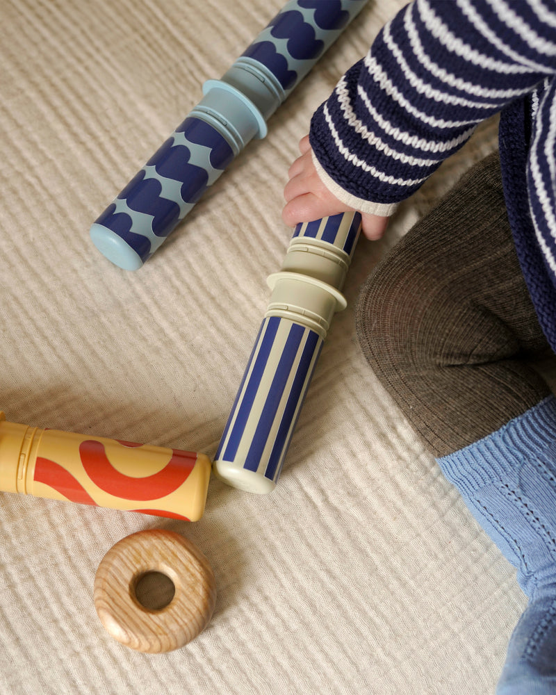 A close-up view of a child's hand holding a dusty green rattle peg from the MODU baby activity set.