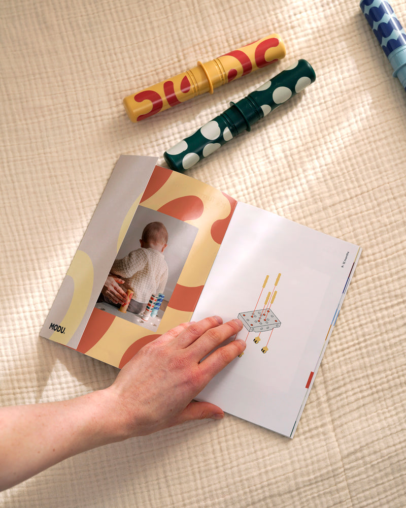 An adult holds open a MODU Activity Guide showing early development play ideas.
