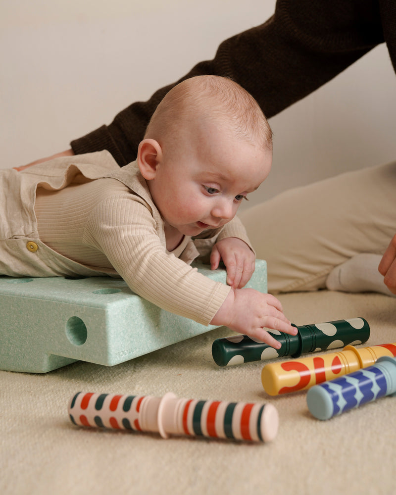 A baby lies on stomach an ocean mint foam block from the MODU Baby Activity Set, reaching toward colorful patterned Rattle Pegs lying on the floor.