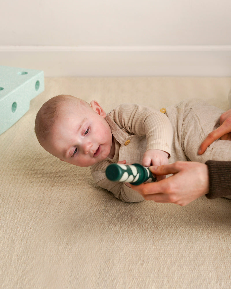 An adult encourages a baby to roll from its back onto its stomach by holding out a green patterned Rattle Peg
