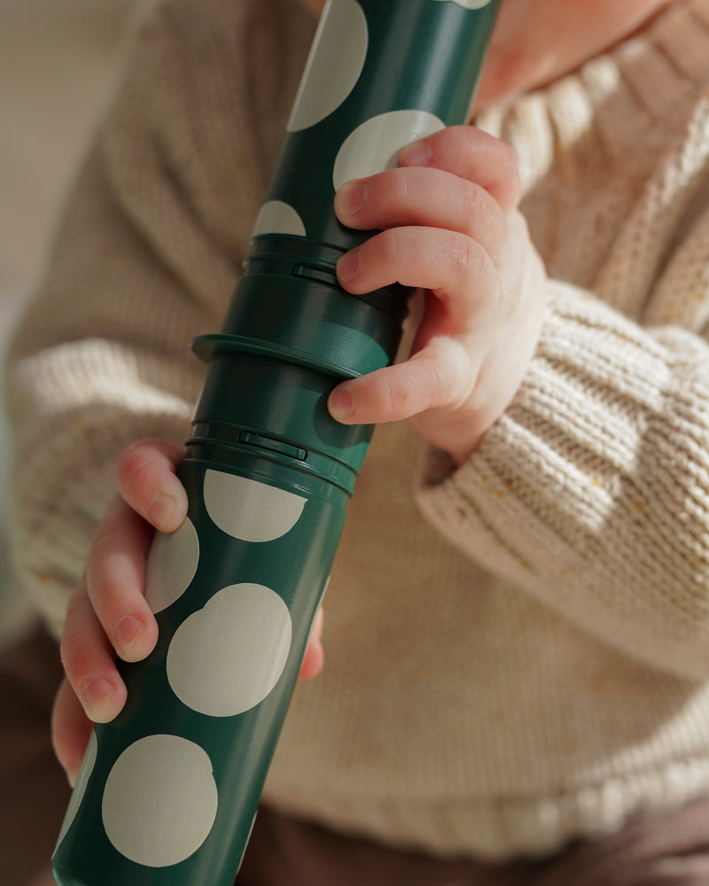 A close-up view of a child's hands holding a forrest green rattle peg from the MODU baby activity set.