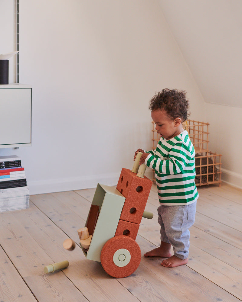 A young toddler tips forward a toy wheelbarrow built from a the 3-in-1 MODU Baby Walker, dropping wooden blocks inside on living room floor.