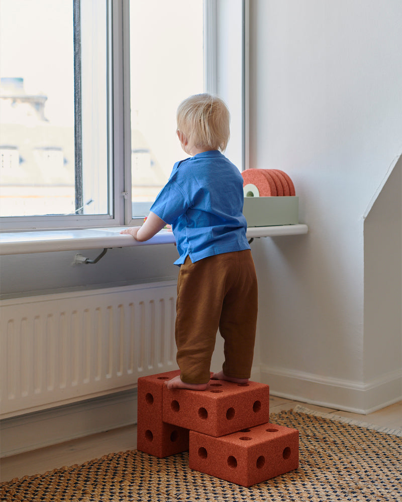 A child looks out a window while standing on a small step stool built from the MODU 3-in-1 Baby Walker.