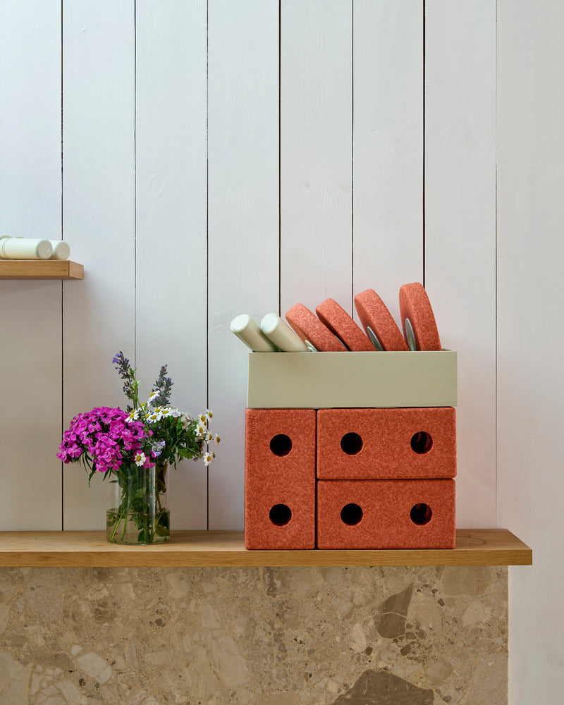 Components of the MODU Baby Walker set in burnt orange and dusty green stored neatly in a box on a shelf next to flowers.