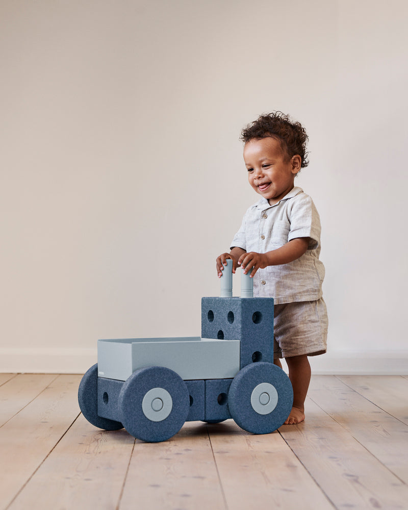 A toddler stands up and holds the handles of a MODU Baby Walker.