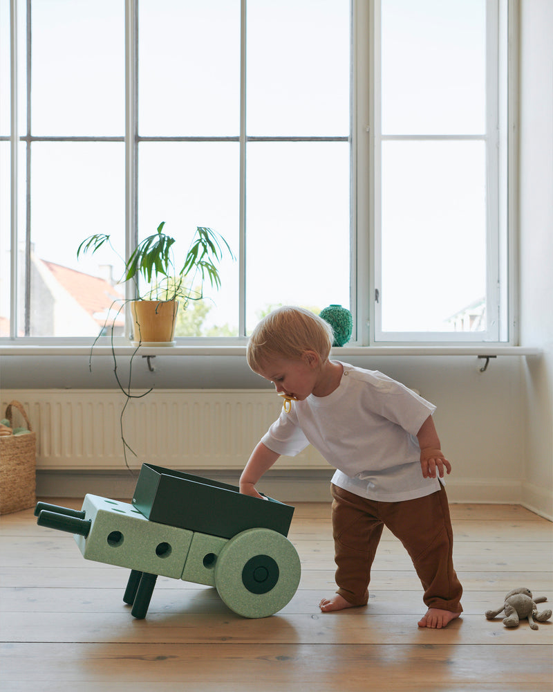 A child reaches into a toy wheelbarrow built from the MODU 3-in-1 Baby Walker that sits on a living room floor.