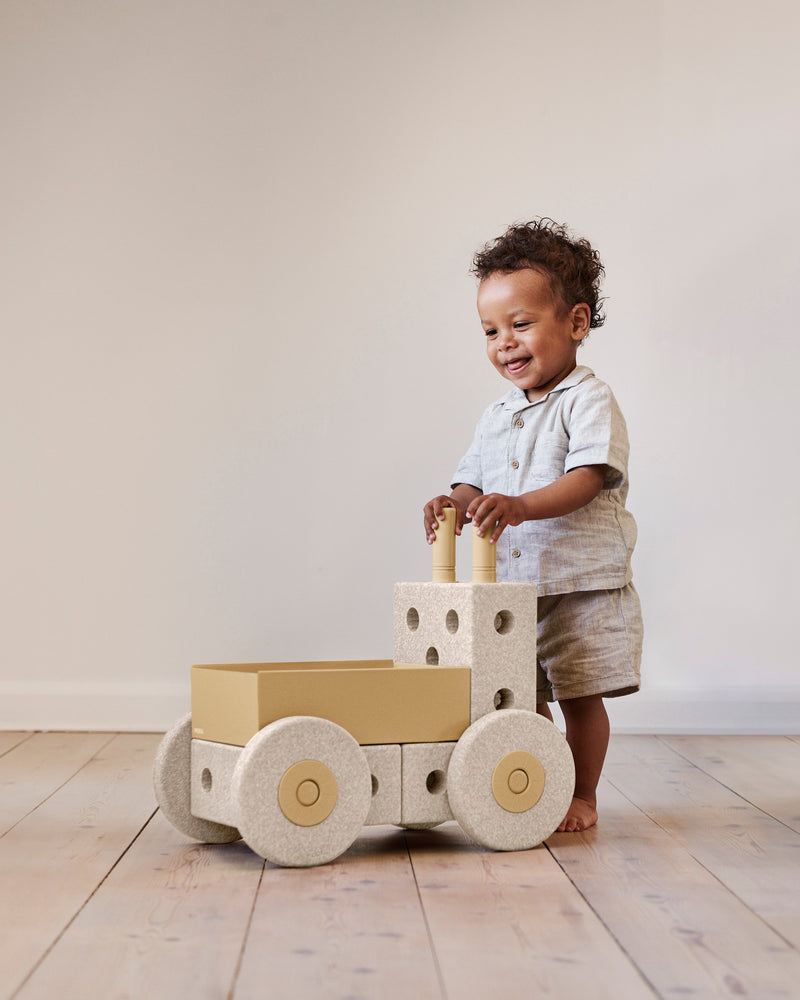 A toddler stands up and holds the handles of a MODU Baby Walker.