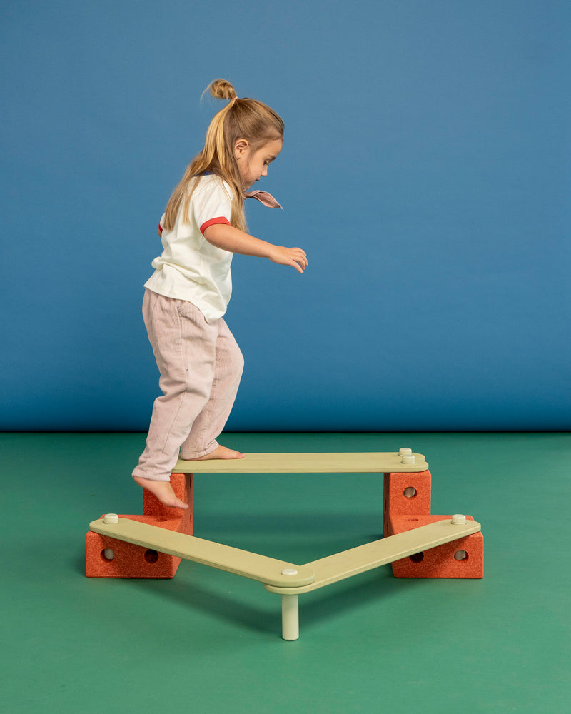 A child carefully steps onto an elevated balance beam circuit built from the MODU Balance Track in burnt orange and dusty green.