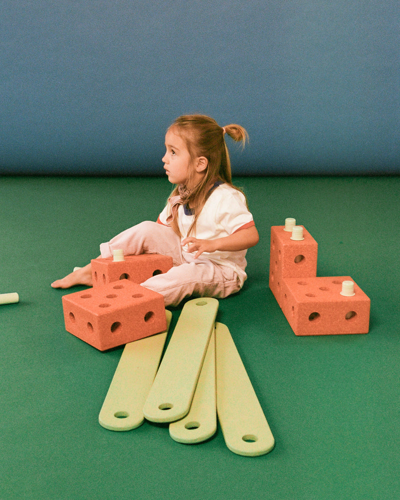 A child sits on a green floor surrounded by burnt orange MODU building blocks and dusty green MODU balance beams.