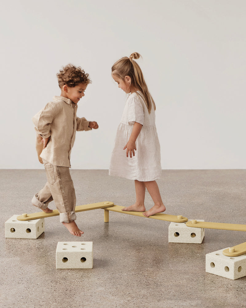 Two children play together, walking and balancing along a MODU Balance Track in sand grey honey yellow.