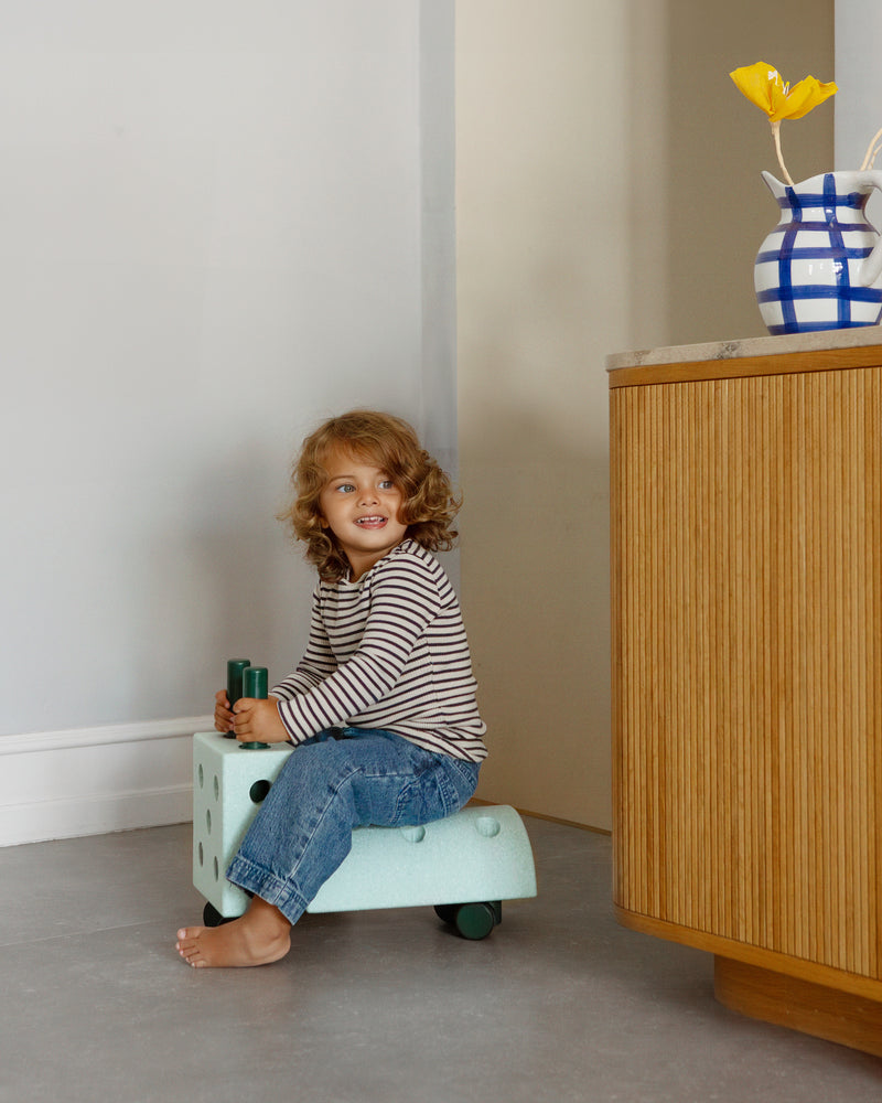 A toddler rides a ride-on toy built from the MODU Curiosity Set in ocean mint across a kitchen floor.