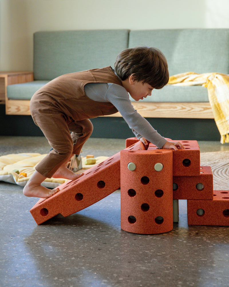 A child climbing up an active play obstacle course with a slide built from the MODU Dreamer Set in burnt orange.