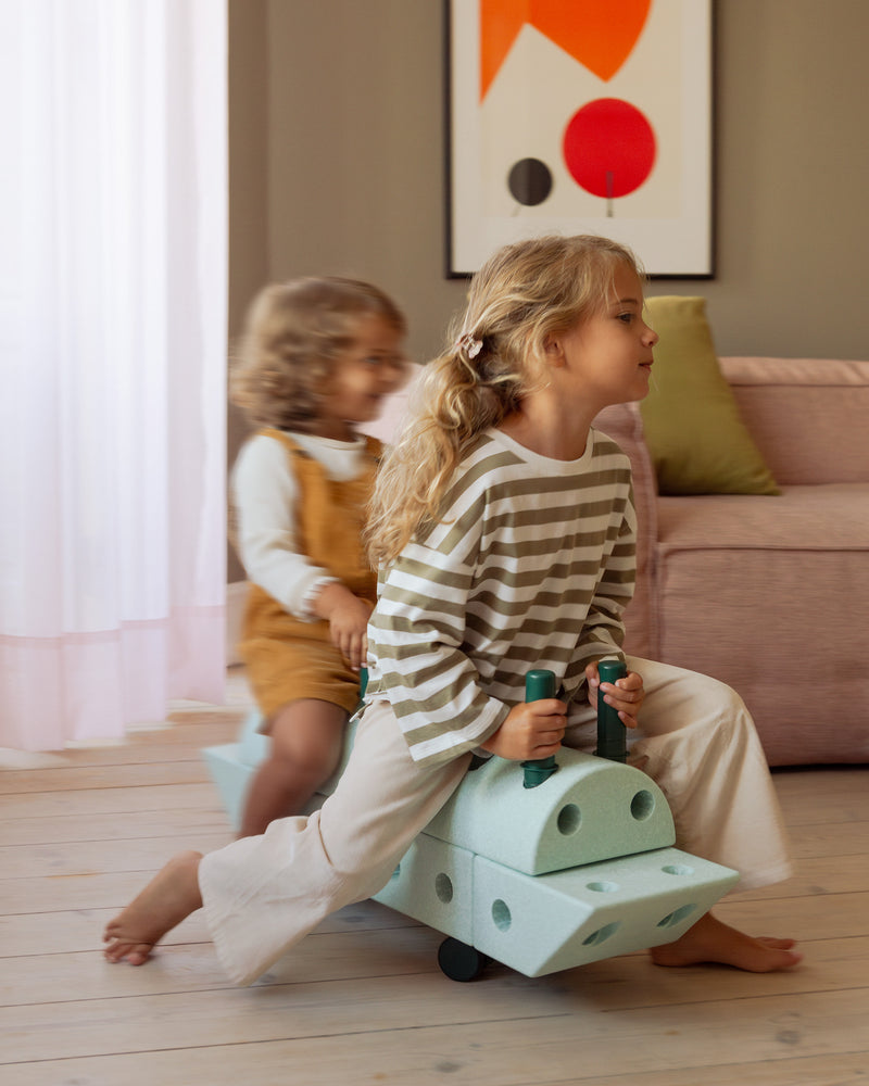 Two children enjoy active play indoors riding a large ride-on toy built from the MODU Dreamer Set.