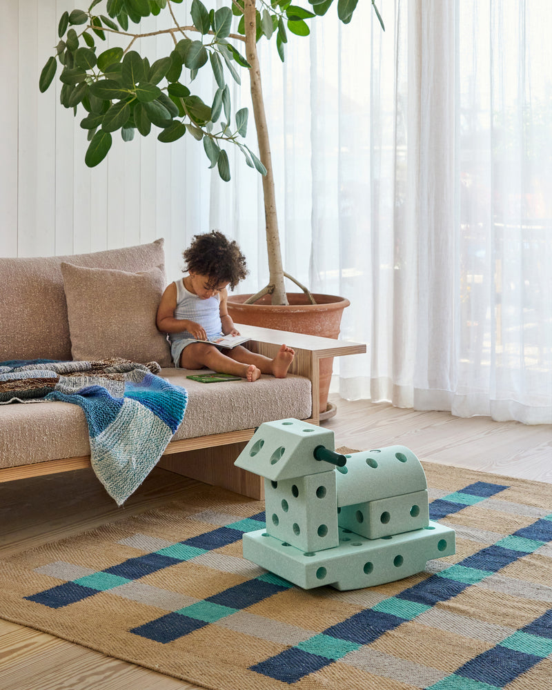 A child sits on a sofa reading a book next to a  modular rocking toy built from the MODU Explorer Set in ocean mint.