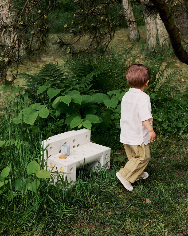 A child walks outdoors near a small bench built from a sand grey MODU Explorer Set with a cup and cookie on it.