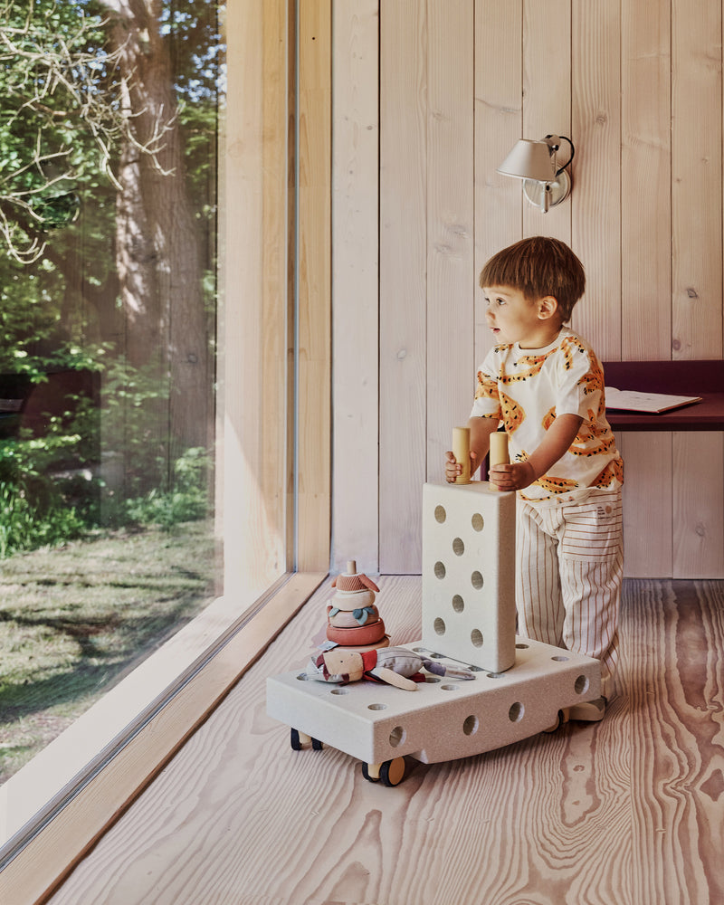 A toddler stands next to a large window pushing toy cart build on caster wheels made from a MODU Explorer Set.