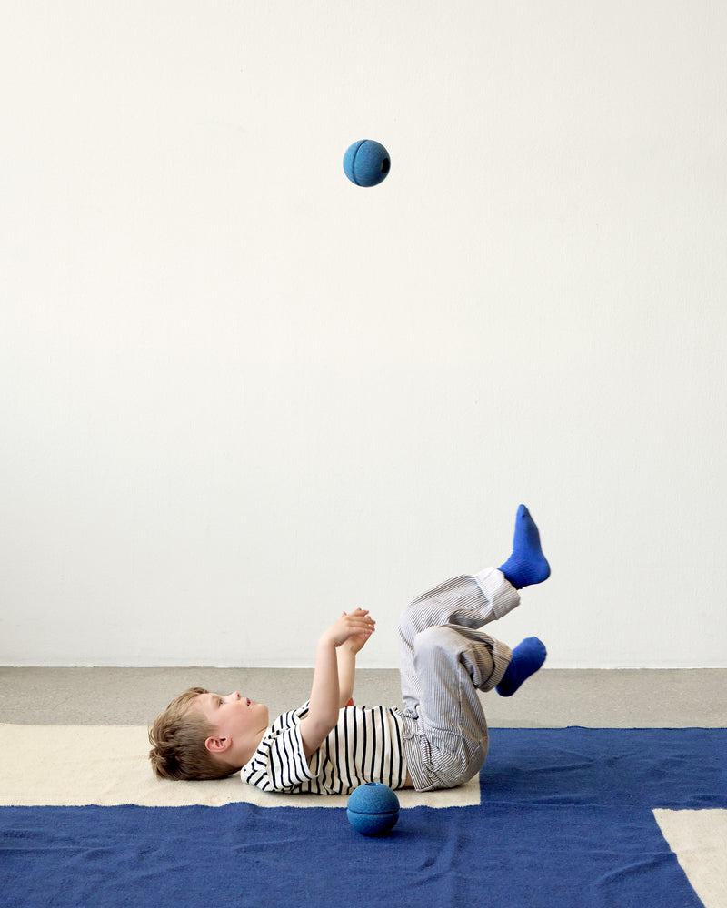 A boy plays with a deep blue MODU Foam Ball on a blue rug, tossing it into the air.
