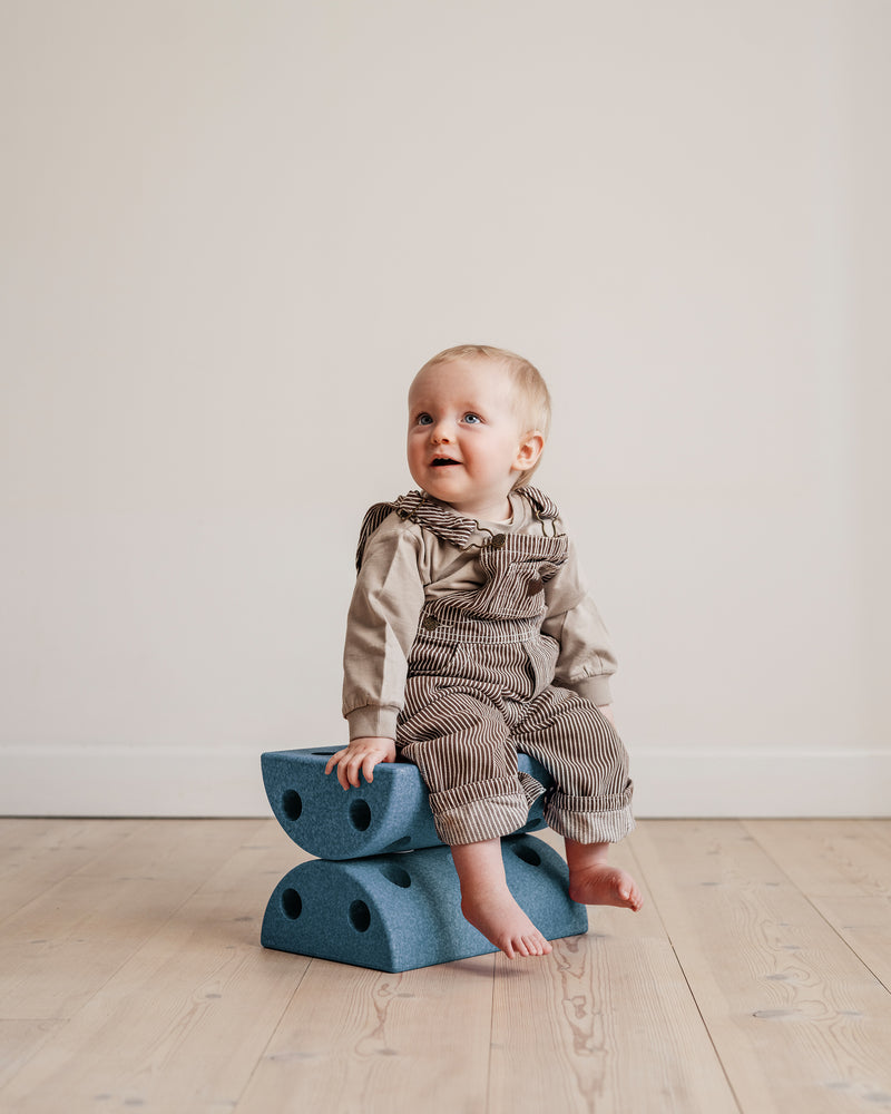 A baby in striped overalls sits safely on a deep blue MODU Kids' Stool.