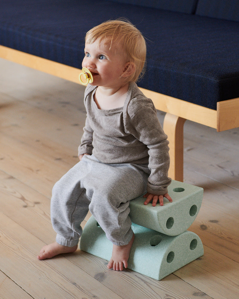 A toddler with a pacifier sits on an ocean mint MODU Kids' Stool in a living room, using it as a low toddler seat.