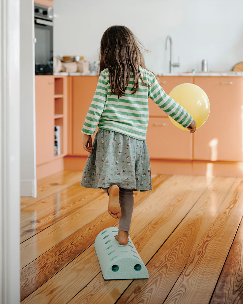 A young girl balancing with a yellow balloon walks along a balance track built from ocean mint MODU Kids' Stool foam blocks.