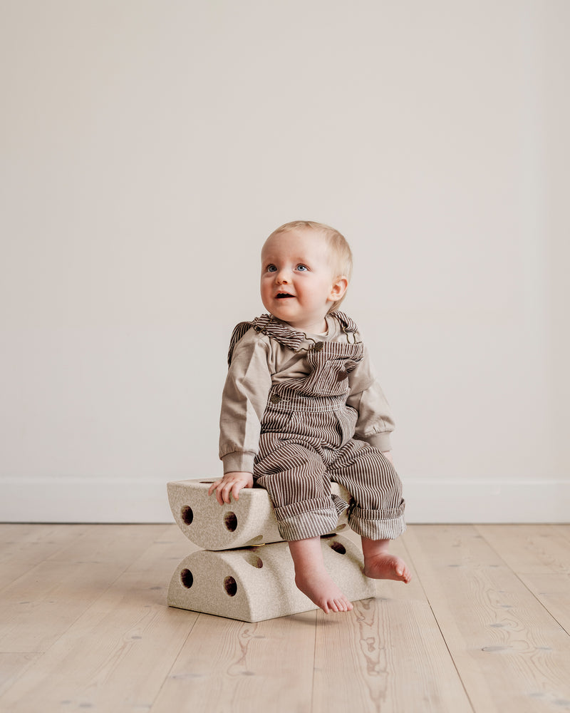 A baby in striped overalls sits on a sand grey MODU Kids' Stool.