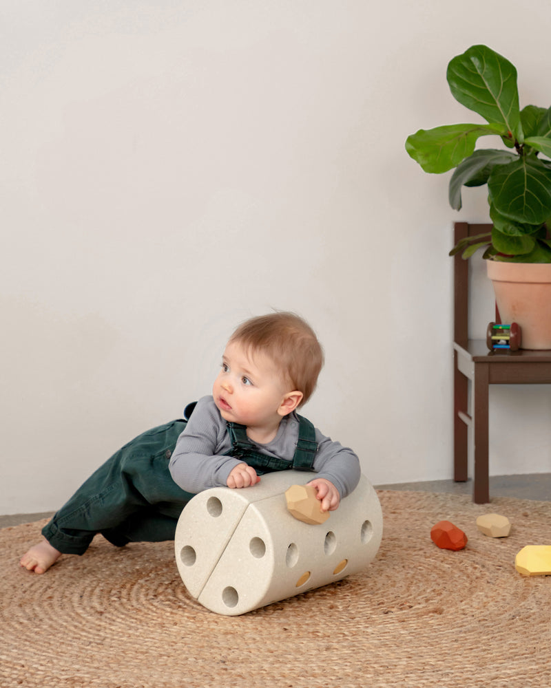 A baby wearing green overalls leans over a sand grey MODU Kids' Stool used as a roller for balance play.
