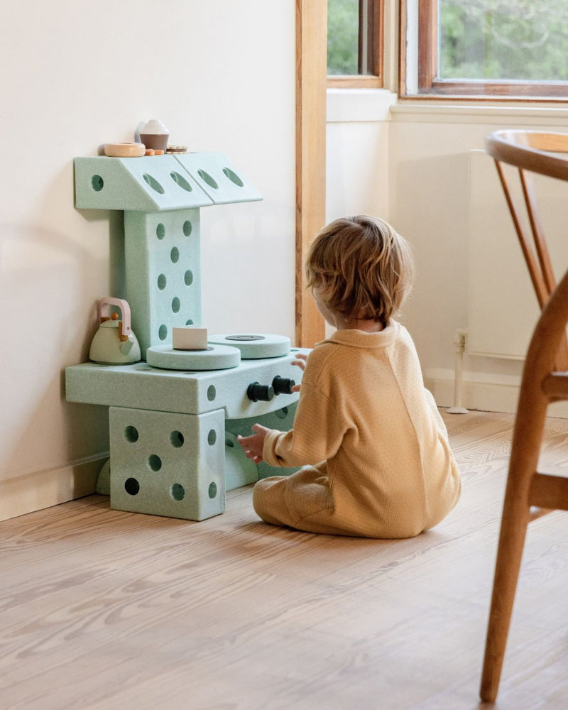 Toddler playing in the living room with MODU play kitchen, pretending to cook and use kitchen utensils. Product color: Ocean Mint Forest Green.