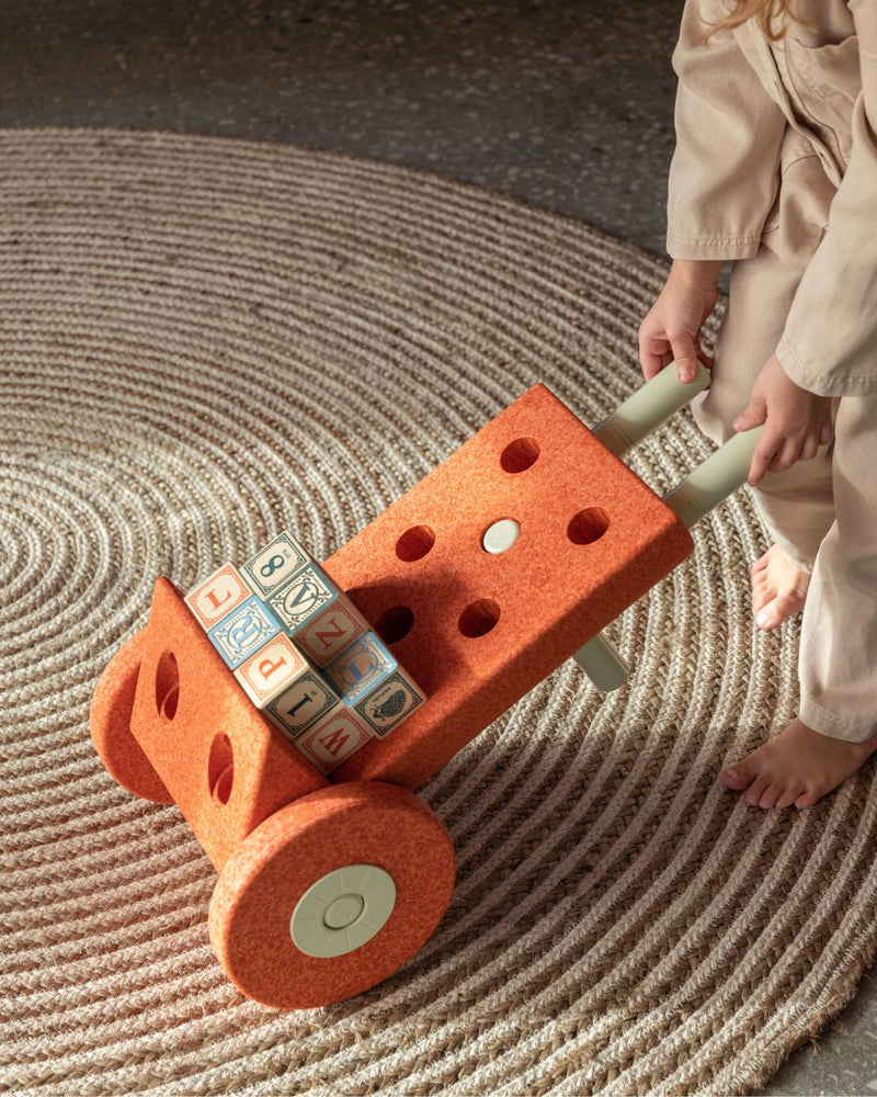 Child pushing MODU wheelbarrow with toys, built from Dreamer Set in Burnt Orange Dusty Green. Encourages gross motor skills.