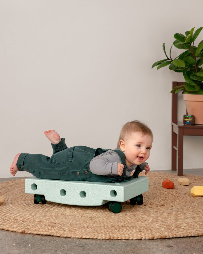 A baby in green overalls smiles during tummy time on an ocean mint MODU Scooter Board placed on a jute rug.
