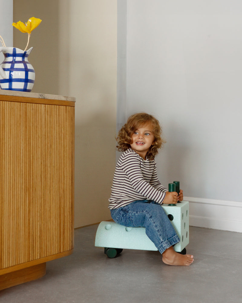 A toddler rides an ocean mint MODU Tiny Ride across kitchen floor.