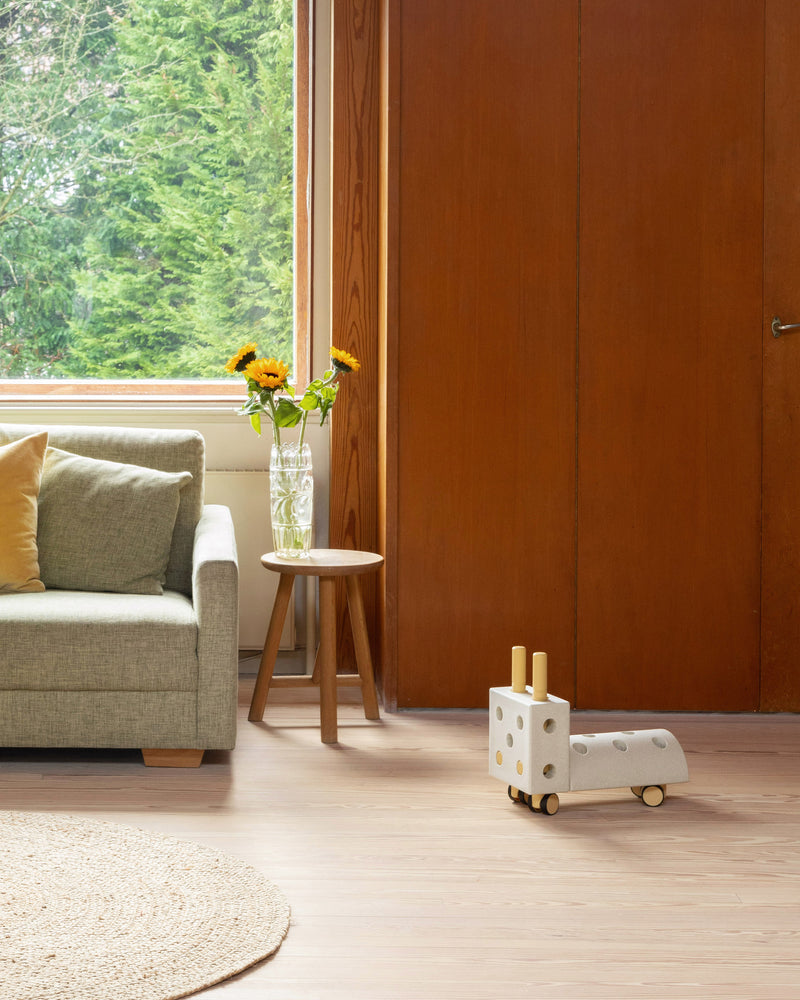 A sand grey and honey yellow MODU Tiny Ride toy ride-on sits on a wooden floor in a living room next to a sofa.