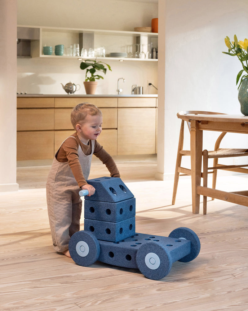 Ein Kind schiebt einen MODU-Lauflernwagen durchs Wohnzimmer und übt Laufen. Hergestellt aus dem Dreamer-Set in Tiefblau und Himmelblau.