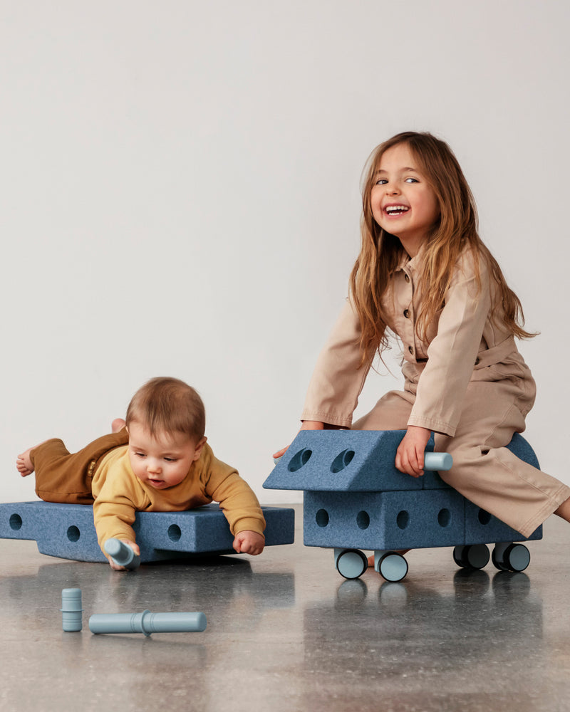 Two children enjoy active play with a ride-on toy and a rocker board build from the MODU Explorer Set.